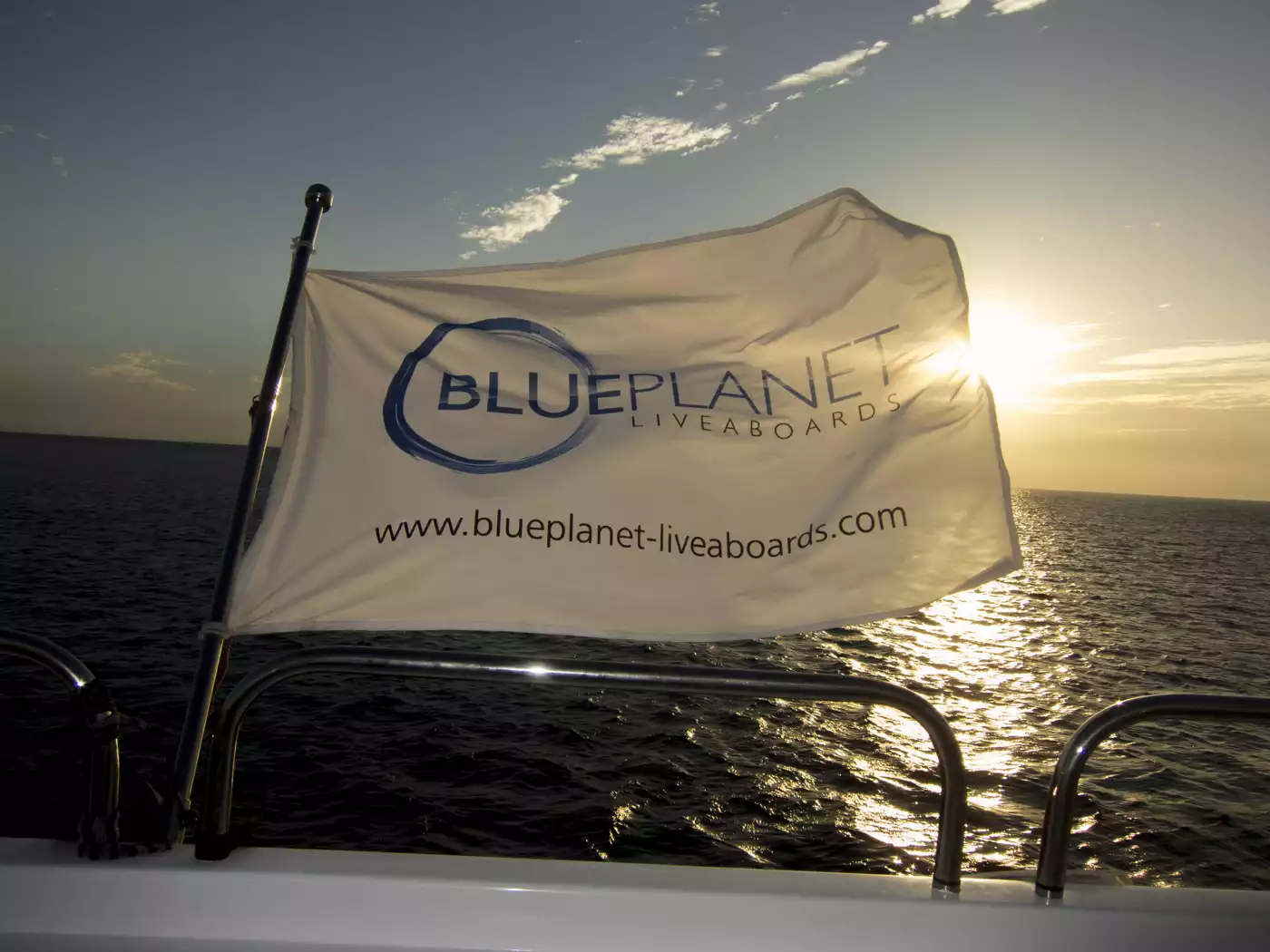 Ein Boot mit einer „Blue Planet Liveaboards“-Flagge ist vor einem Sonnenuntergang über dem Meer zu sehen. Die Sonne wird teilweise von der Flagge verdeckt und wirft einen warmen Schein auf das Wasser. Auf der Flagge sind das Firmenlogo und die URL zu sehen.