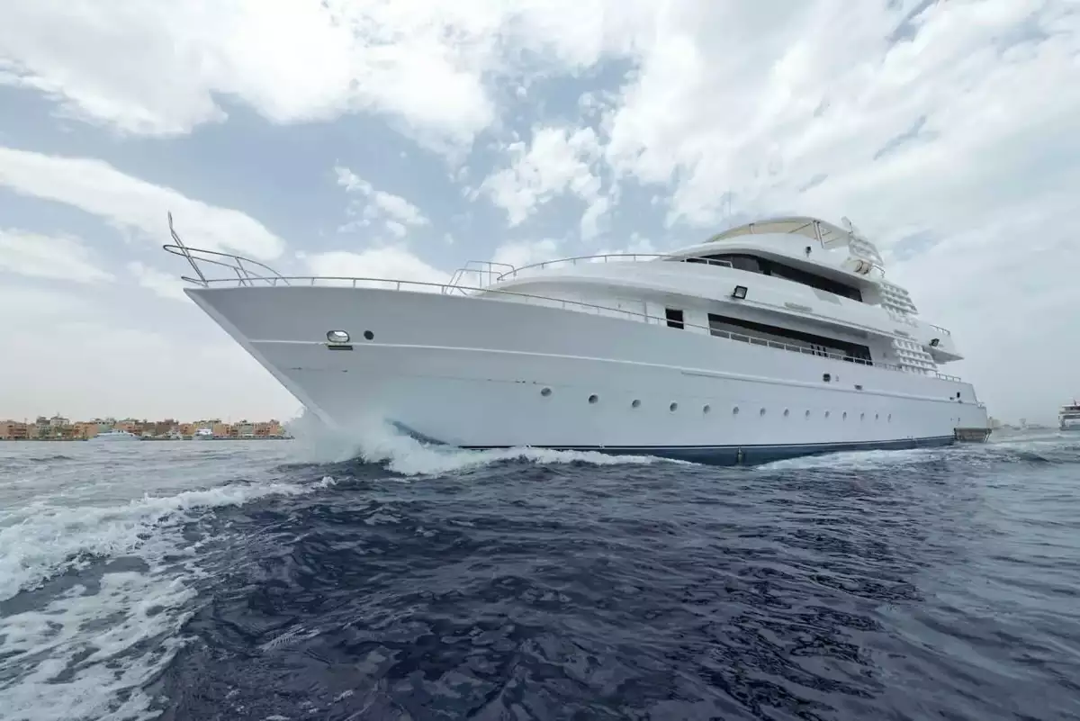 Eine große, weiße Luxusyacht kreuzt auf offener See unter einem teilweise bewölkten Himmel. Die Yacht hat mehrere Decks und erzeugt Wellen, während sie durch das Wasser gleitet. Im Hintergrund ist eine entfernte Küstenlinie sichtbar.