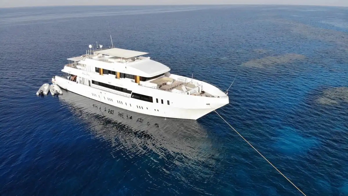 Eine große weiße Yacht ankert im klaren blauen Wasser. Das Schiff hat mehrere Decks, ein elegantes Design und eine Reihe kleinerer Boote, die an seiner Seite befestigt sind. Das Meer ist ruhig, Sonnenflecken spiegeln sich auf der Oberfläche. Im Hintergrund ist kein Land zu sehen.