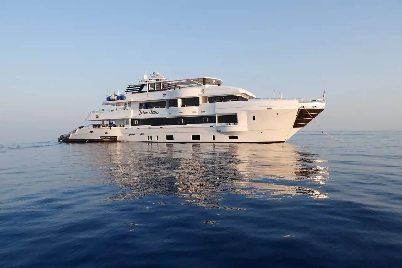 Eine große Luxusyacht namens „Blue Storm“ schwimmt ruhig auf dem klaren blauen Ozean. Der Himmel ist wolkenlos und die Wasseroberfläche ist ruhig und spiegelt das weiße Äußere und die mehreren Decks der Yacht wider.