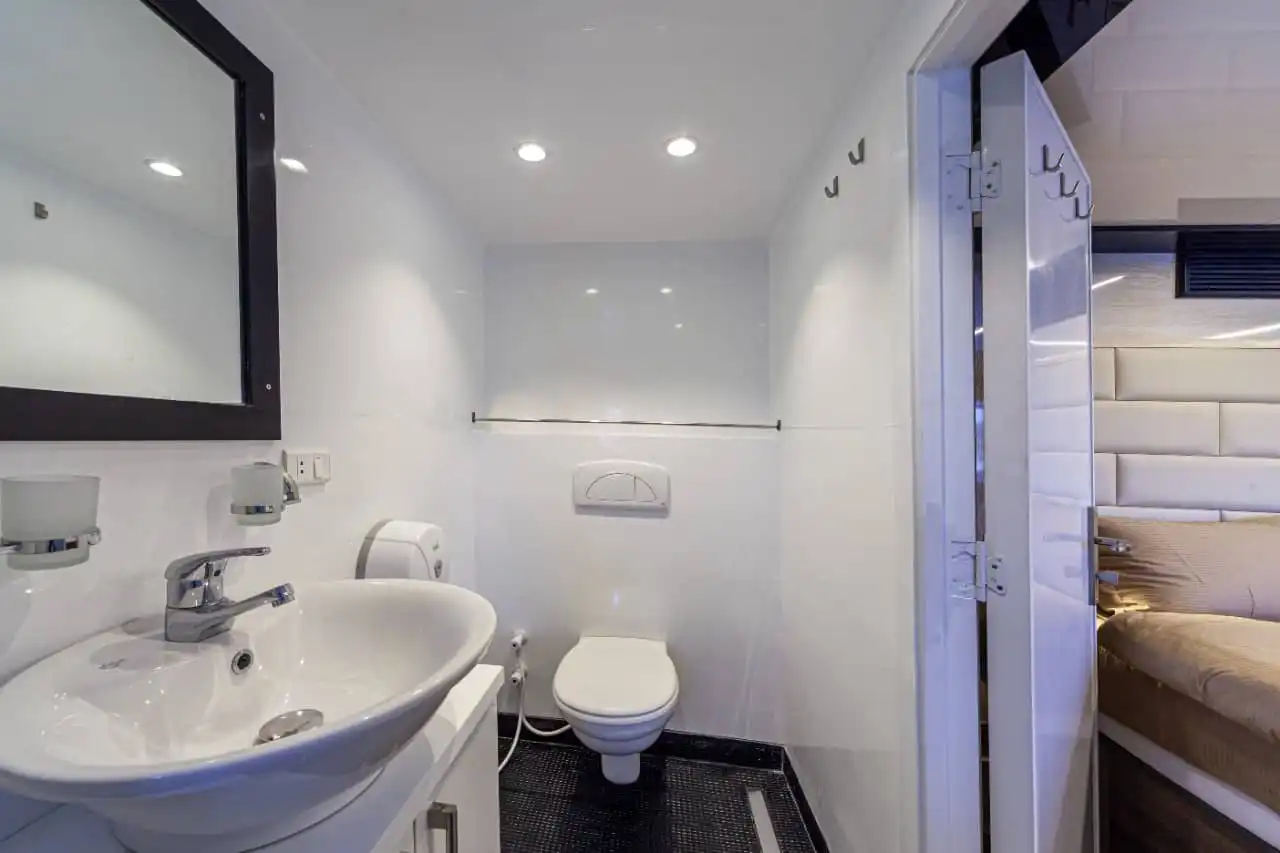 Ein kleines, modernes Badezimmer mit weißen Wänden und eleganten Armaturen. Es verfügt über ein wandmontiertes Waschbecken mit einem Spiegel darüber, eine Toilette und Deckenleuchten. Der Boden ist dunkel gefärbt und auf der rechten Seite befindet sich eine teilweise geöffnete Tür.