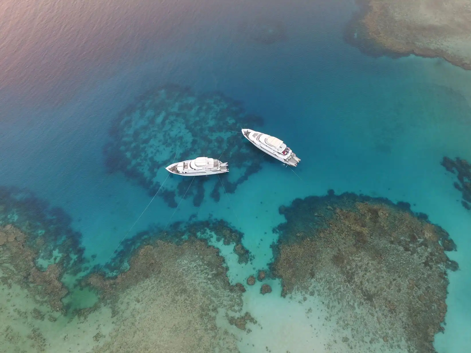 Luftaufnahme von zwei Yachten, die in klarem, türkisfarbenem Wasser vor Anker liegen und von Korallenriffen umgeben sind. Das Bild hebt den Kontrast zwischen den tiefen und flachen Bereichen des Ozeans hervor, wobei unter der Oberfläche Korallenformationen sichtbar sind.
