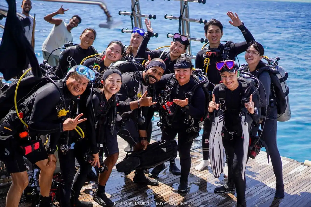 Unser Team aus elf Personen in Taucherausrüstung lächelt und posiert auf einem Bootsdeck am Meer. Sie wirken fröhlich und bereit für ein Tauchabenteuer, im Hintergrund ist das blaue Meer zu sehen.